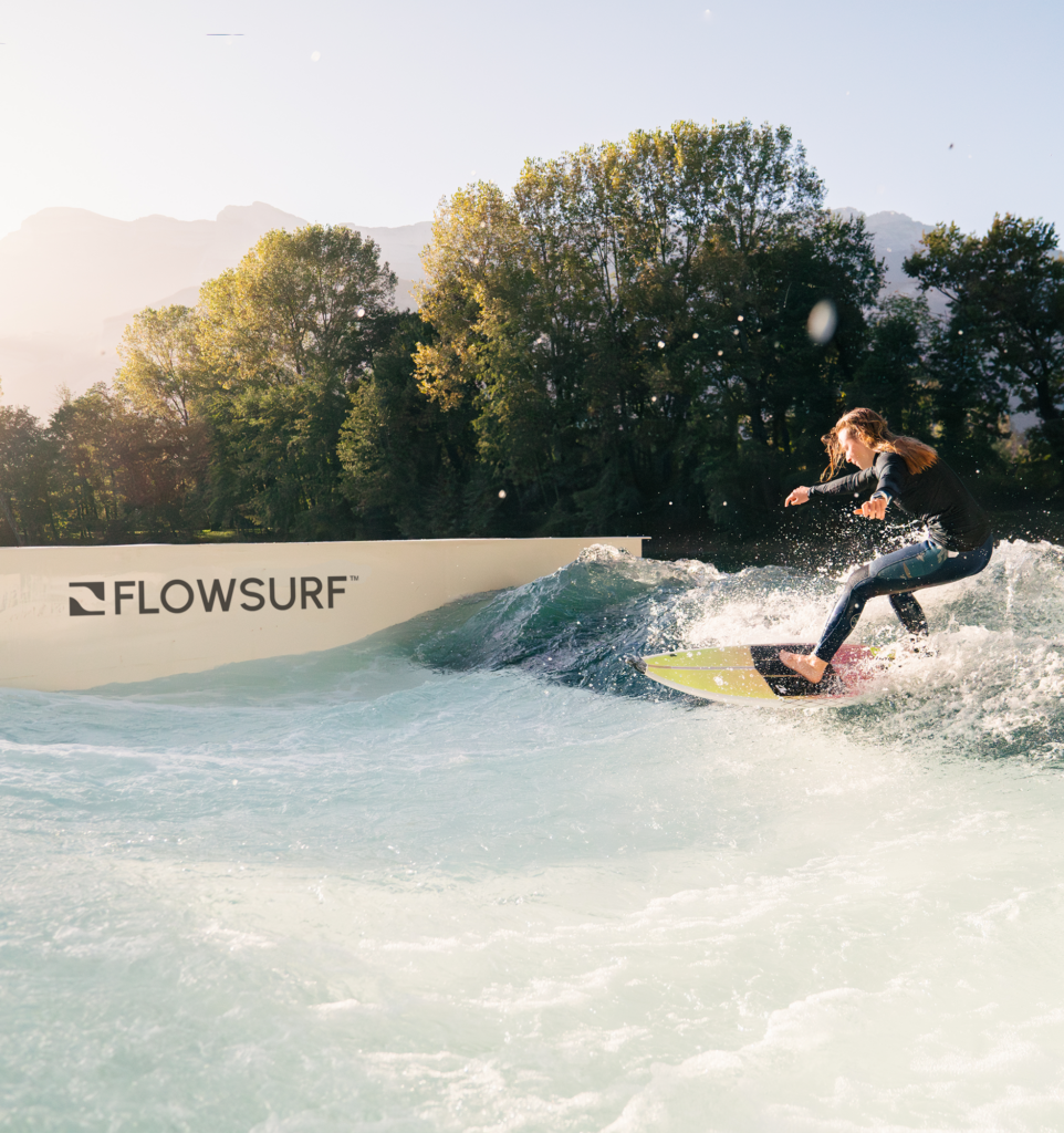 A person in a wetsuit surfs on an artificial wave at a facility with FLOWSURF branding, surrounded by trees, under a clear sky with sunlight streaming through.