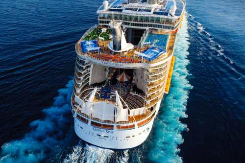 Aerial view of the cruise ship Oasis of the Seas sailing on blue ocean water, showing open decks with pools, green spaces, and recreational areas, leaving a white wake behind.
