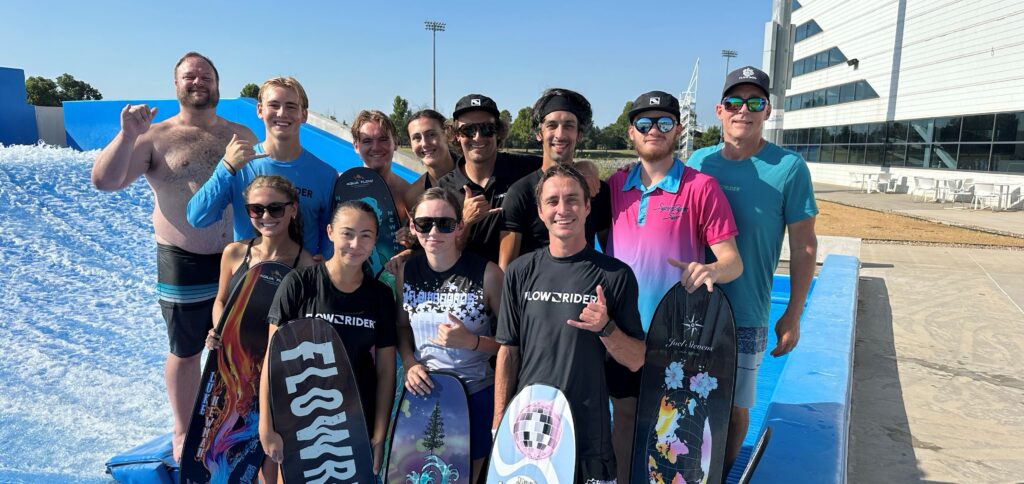 A group of thirteen smiling people, some holding flowboards, pose together in swimwear and casual clothes in front of an outdoor artificial wave surfing pool during a sunny Flow Tour event.