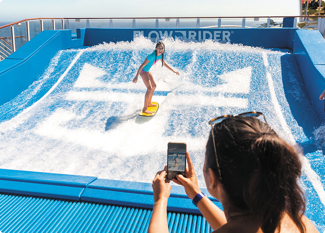 A person rides a wave on a FlowRider surf simulator while another person in the foreground takes a photo with a smartphone. The scene is outdoors on a sunny day, possibly on a cruise ship.