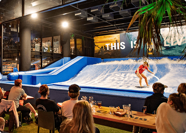 A group of people sit at tables watching a person surf on an indoor artificial wave. The scene is lively, with drinks on the tables and a large sign in the background reading THIS IS.