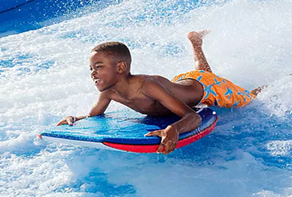 A young boy in orange swim trunks rides a blue bodyboard on a wave at a water park, with water splashing around him as he smiles and enjoys the activity.