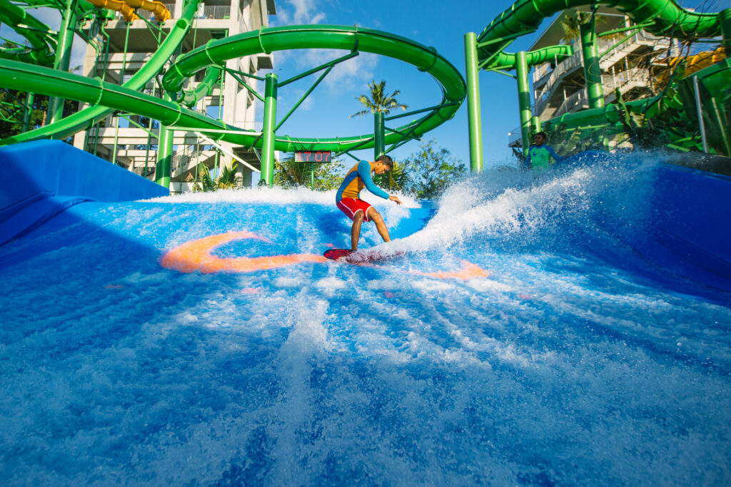A person in swimwear rides an artificial wave on a blue surfing simulator at a water park, surrounded by green water slides and clear blue sky in the background.