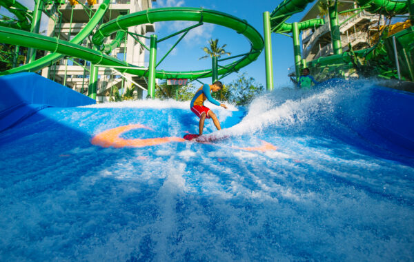 A person in swimwear rides an artificial wave on a blue surfing simulator at a water park, surrounded by green water slides and clear blue sky in the background.