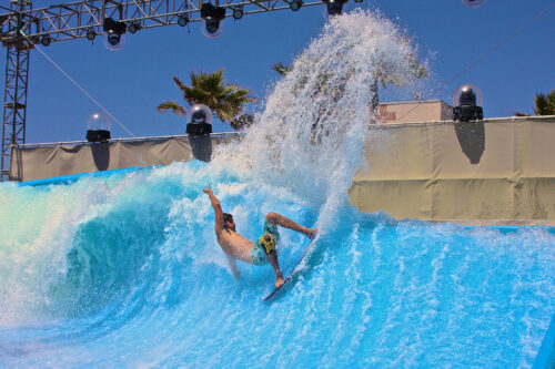 A surfer rides a wave on an artificial surfing pool, kicking up a large spray of water under a clear blue sky with palm trees and stage lights in the background.