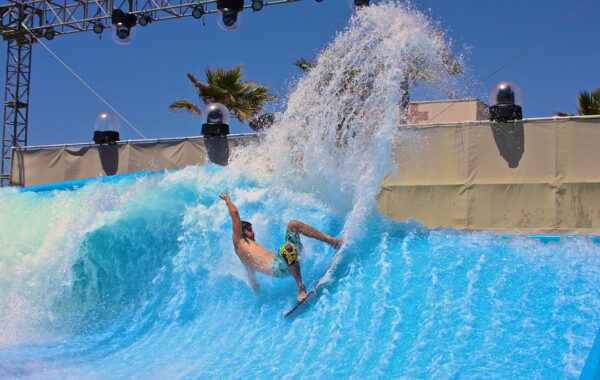 A surfer rides a wave on an artificial surfing pool, kicking up a large spray of water under a clear blue sky with palm trees and stage lights in the background.
