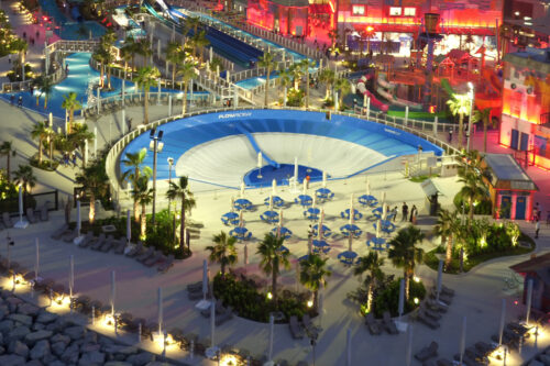 An illuminated water park at night featuring a large circular wave simulator, blue sun umbrellas, lounge chairs, palm trees, and various water slides in the background.