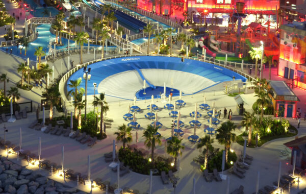 An illuminated water park at night featuring a large circular wave simulator, blue sun umbrellas, lounge chairs, palm trees, and various water slides in the background.