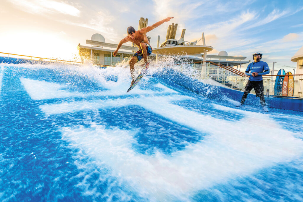 A man surfs on an artificial wave machine, catching air above the water, while another person watches nearby. The scene takes place outdoors, possibly on a cruise ship, under a bright, blue sky.