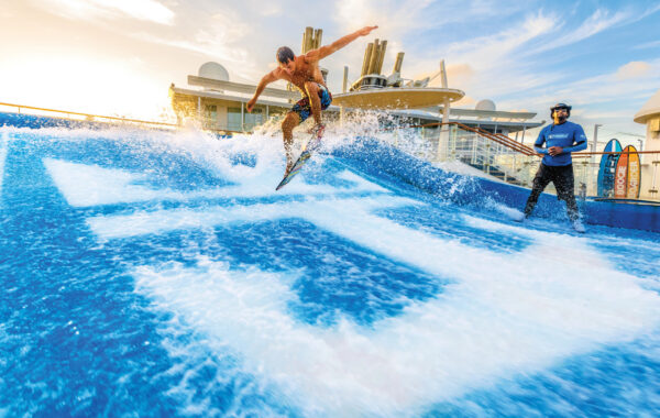 A man surfs on an artificial wave machine, catching air above the water, while another person watches nearby. The scene takes place outdoors, possibly on a cruise ship, under a bright, blue sky.