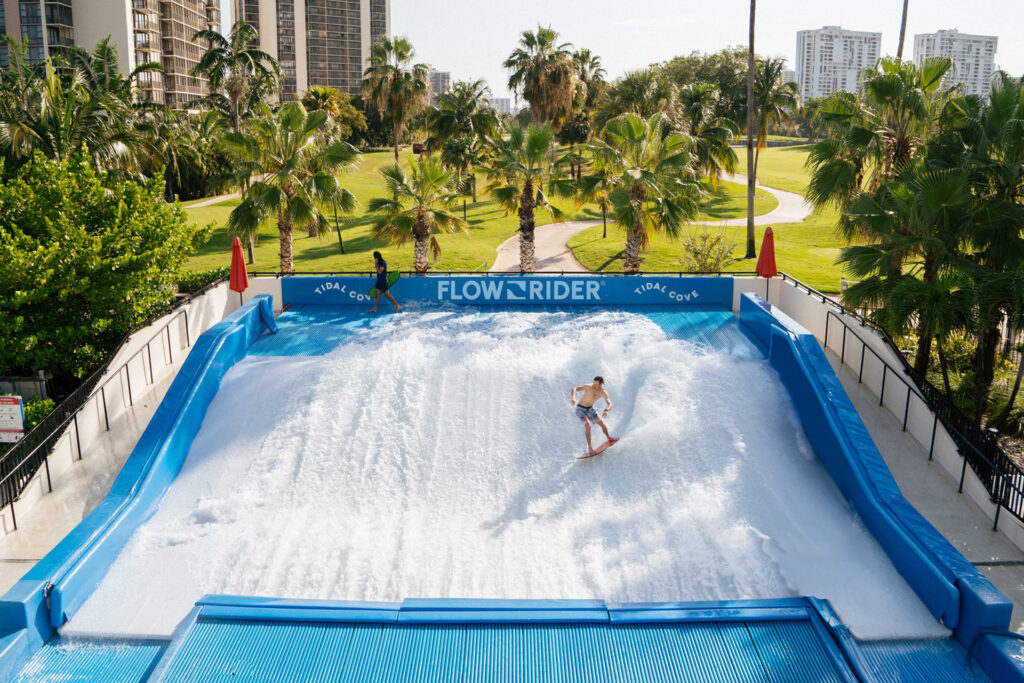 A person surfs on an artificial wave at a FlowRider attraction surrounded by palm trees, with tall buildings and a grassy park visible in the background under a clear sky.