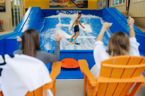 A man surfs on an indoor wave simulator while two people in orange chairs cheer him on with raised arms. The scene is lively and colorful, with water splashing around the surfer.