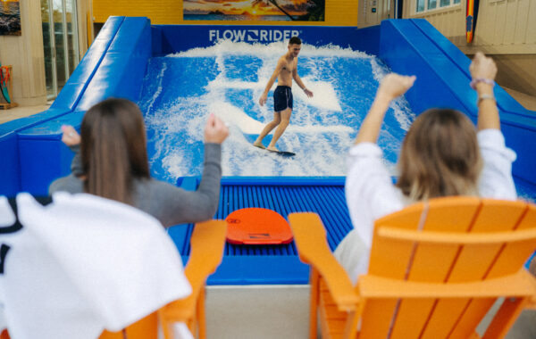 A man surfs on an indoor wave simulator while two people in orange chairs cheer him on with raised arms. The scene is lively and colorful, with water splashing around the surfer.