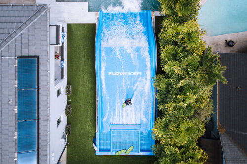A person surfs on a blue FlowRider wave simulator surrounded by trees, grass, and nearby buildings, seen from an overhead aerial view.