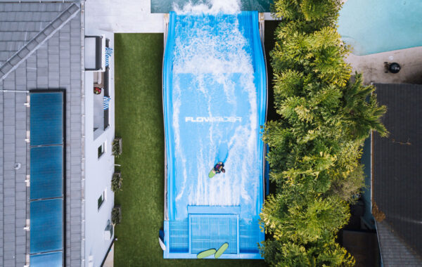 A person surfs on a blue FlowRider wave simulator surrounded by trees, grass, and nearby buildings, seen from an overhead aerial view.