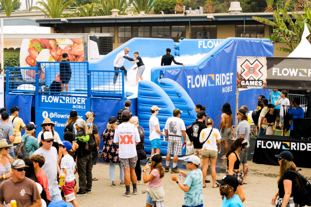A crowd watches as people surf on an artificial wave machine at an outdoor FlowRider attraction, with palm trees and event tents in the background. Multiple people are queuing and enjoying the lively atmosphere.