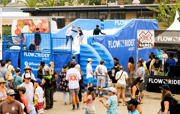 A crowd watches as people surf on an artificial wave machine at an outdoor FlowRider attraction, with palm trees and event tents in the background. Multiple people are queuing and enjoying the lively atmosphere.