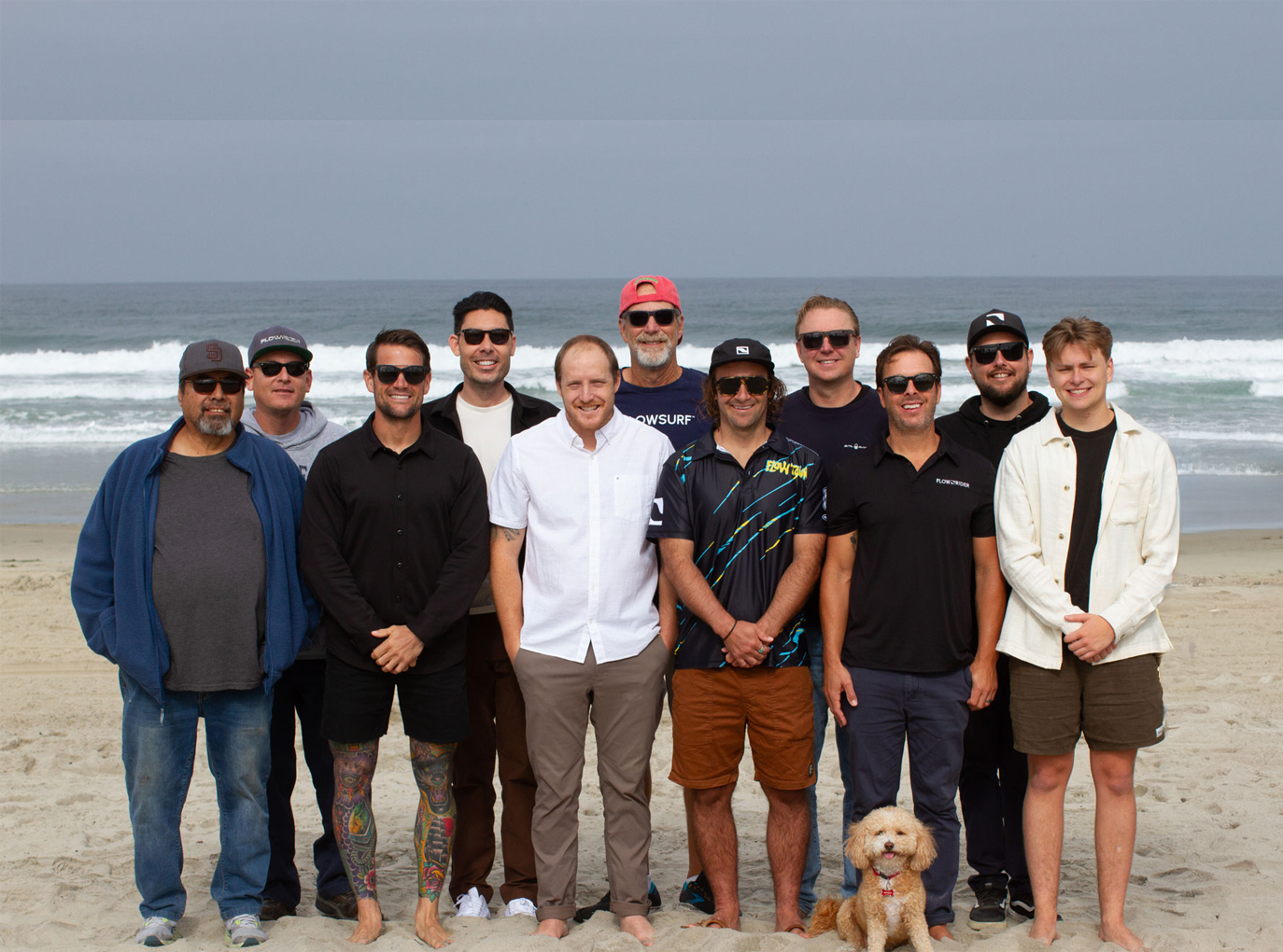 Twelve men and a small dog pose together on a sandy beach with waves in the background. The group is smiling, dressed casually, and the weather appears mild and overcast.