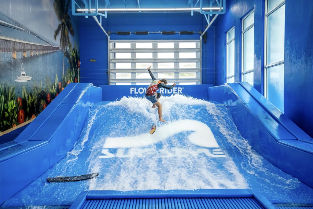 A person surfs on an indoor artificial wave machine during the Flow Tour, surrounded by blue walls and windows. A mural of a beach and a bridge is visible on the left wall, with a surfboard lying on the floor nearby.