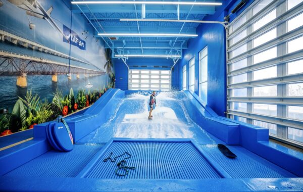 A child rides a wave on an indoor surf simulator during a Flow Tour event in a bright, blue room with a beach mural on the wall. Large windows let in natural light, and surfboards and gear are visible on the side.