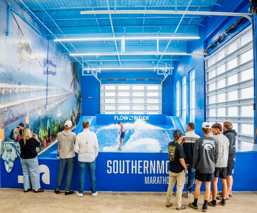 A group of people watch someone surfing on an indoor FlowRider wave simulator during a Flow Tour event, in a bright blue room with large windows and a mural of a plane and tropical scene on the wall.