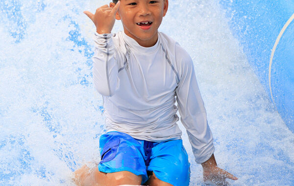 A smiling boy in a white rash guard and blue swim trunks rides a red bodyboard on a wave, making a shaka hand sign with his right hand as water splashes around him.