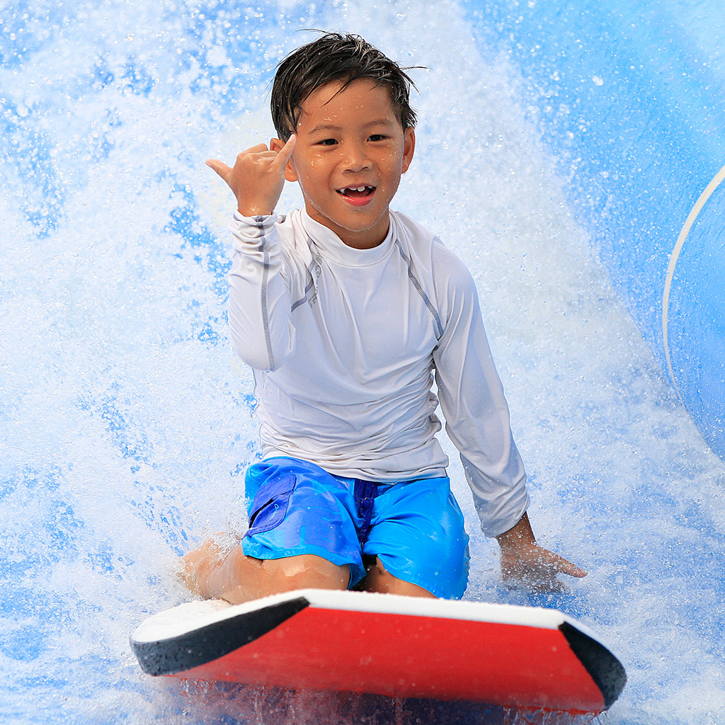 A smiling boy in a white rash guard and blue swim trunks rides a red bodyboard on a wave, making a shaka hand sign with his right hand as water splashes around him.