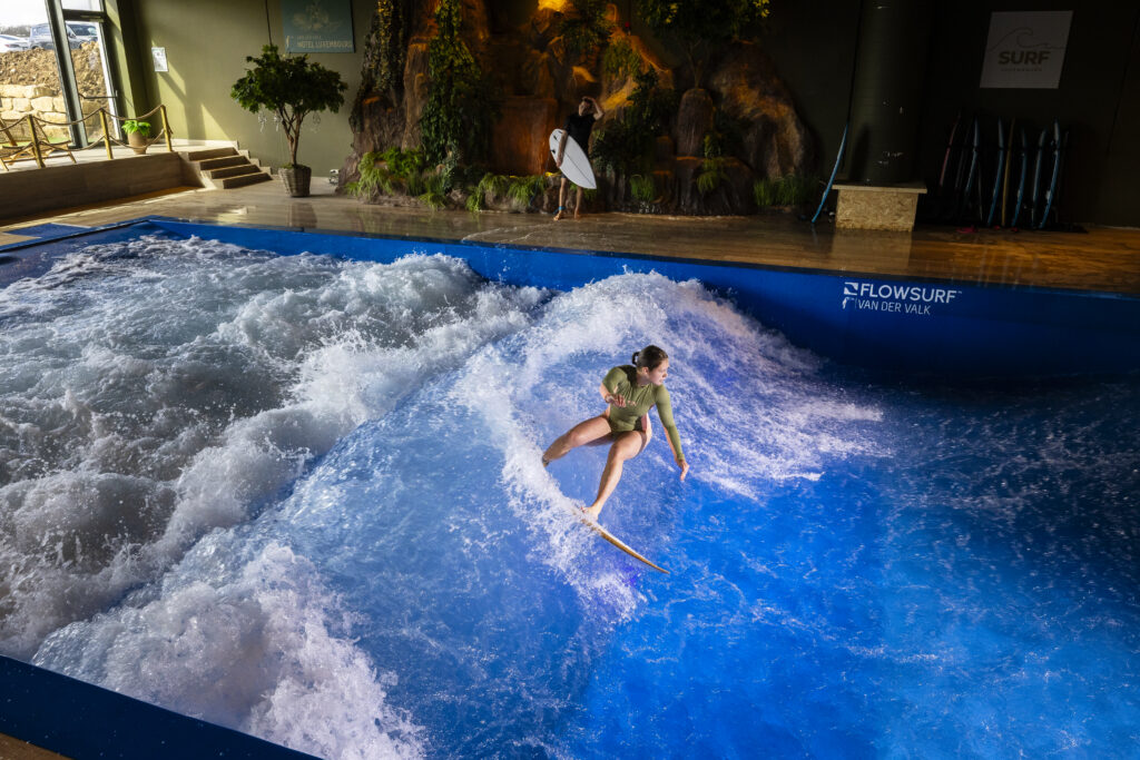 A person surfs on an indoor artificial wave, balancing on a board as water rushes around them. Another person holding a surfboard stands in the background near a rocky, tropical-themed wall.