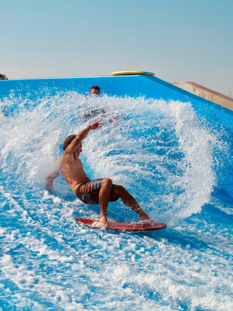 A man in swim trunks rides a wave on a red surfboard, leaning sharply and spraying water, while another person stands in the background on a blue artificial wave pool.