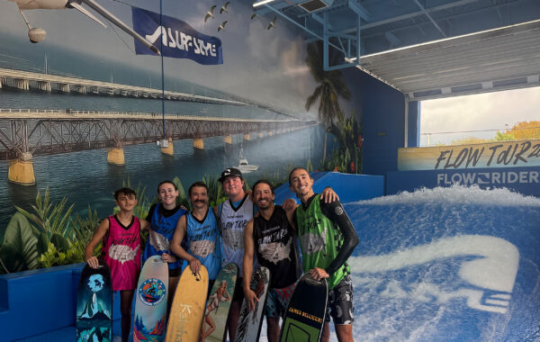 Six smiling people in wet gear hold bodyboards inside a surf park, standing in front of a wave machine with a mural of a bridge and ocean on the wall behind them. A brightly colored FLOW TOUR banner hangs above the wave, highlighting the excitement and competitive spirit of the Flow Tour event.