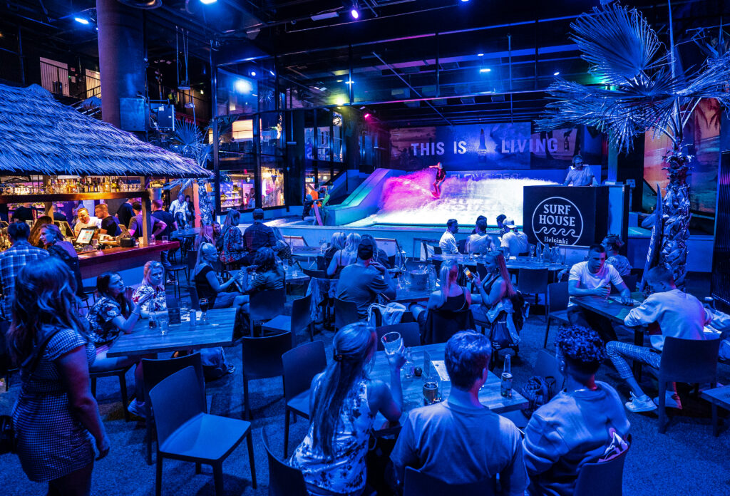 People dine and socialize in a vibrant, tropical-themed indoor venue with blue lighting. In the background, someone surfs on the FlowSurf artificial wave under a sign that reads THIS IS LIVING. A tiki-style bar is on the left.
