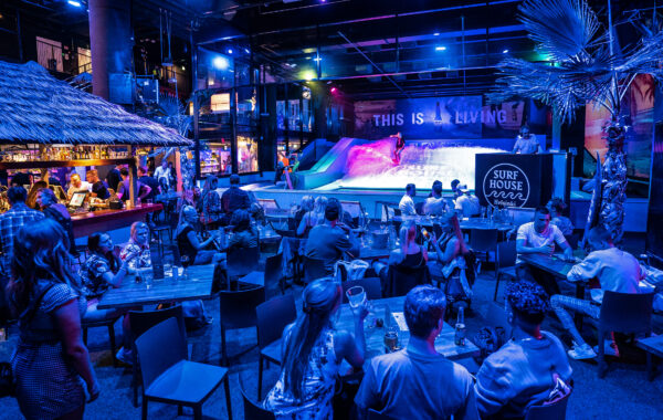 People dine and socialize in a vibrant, tropical-themed indoor venue with blue lighting. In the background, someone surfs on the FlowSurf artificial wave under a sign that reads THIS IS LIVING. A tiki-style bar is on the left.