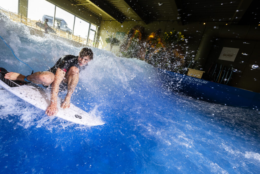 A young man in a wetsuit surfs on an artificial wave indoors during a Flow Tour event, crouching low on his white surfboard as water splashes around him under bright lights.