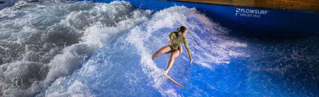 A person in a green wetsuit surfs on an artificial indoor wave during the Flow Tour, surrounded by splashing water at an indoor surfing facility marked FLOWSURF and VANDER VALK on the blue wall.
