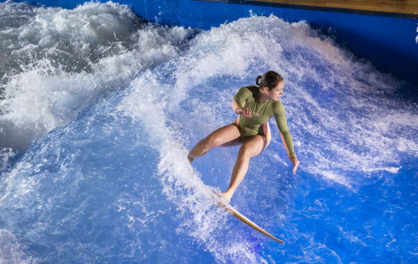 A person in a green wetsuit surfs on an artificial indoor wave during the Flow Tour, surrounded by splashing water at an indoor surfing facility marked FLOWSURF and VANDER VALK on the blue wall.