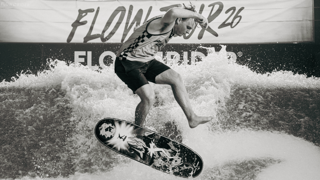 A surfer performs a trick on an indoor wave, balancing on a board with water splashing around. A Flow Tour banner is visible in the background, capturing the excitement of this black and white action shot.