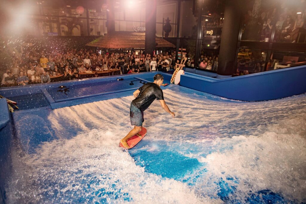 A man surfs on a FlowSurf indoor artificial wave surrounded by blue barriers, while a large crowd watches from behind the glass in the background. The scene is brightly lit and energetic.