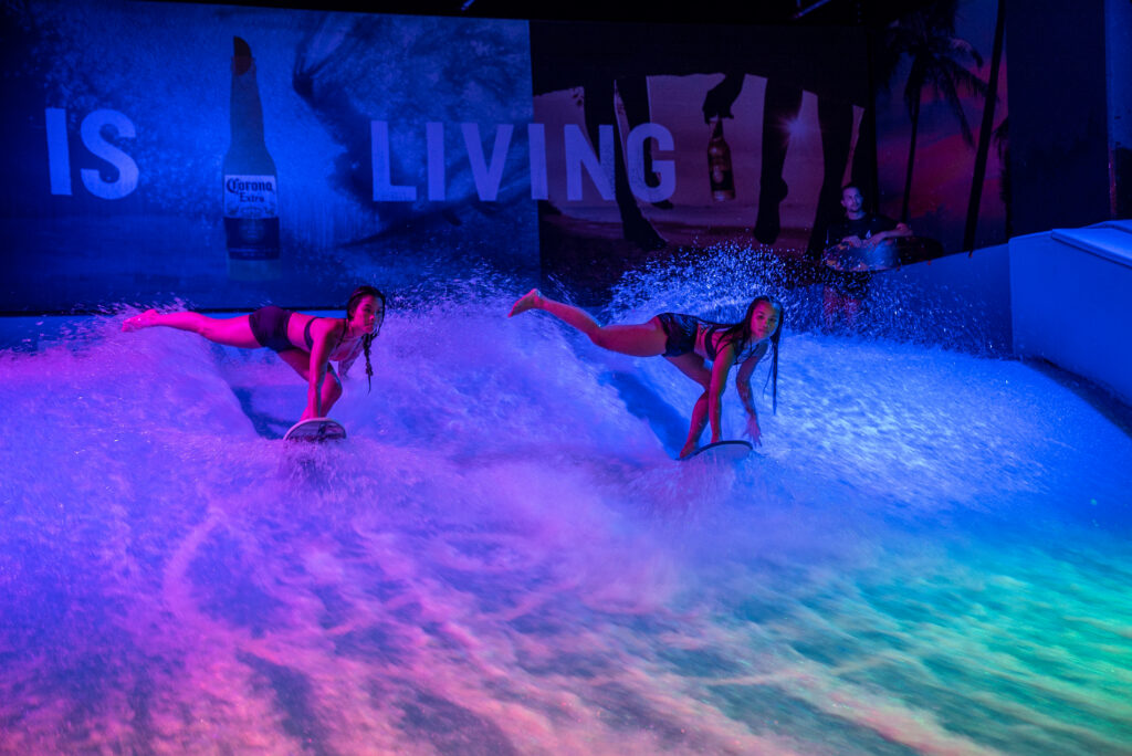 Two women ride the FlowSurf indoor wave under vibrant lights, splashing water as they balance. A man watches nearby. A large sign displaying "IS LIVING" and a Corona beer bottle decorates the wall in the background.