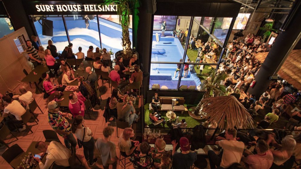 A large crowd gathers at Surf House Helsinki, watching FlowSurf sessions through glass windows. People sit at tables under palm trees and tiki-style decor, enjoying food and drinks in a lively, tropical-themed atmosphere.
