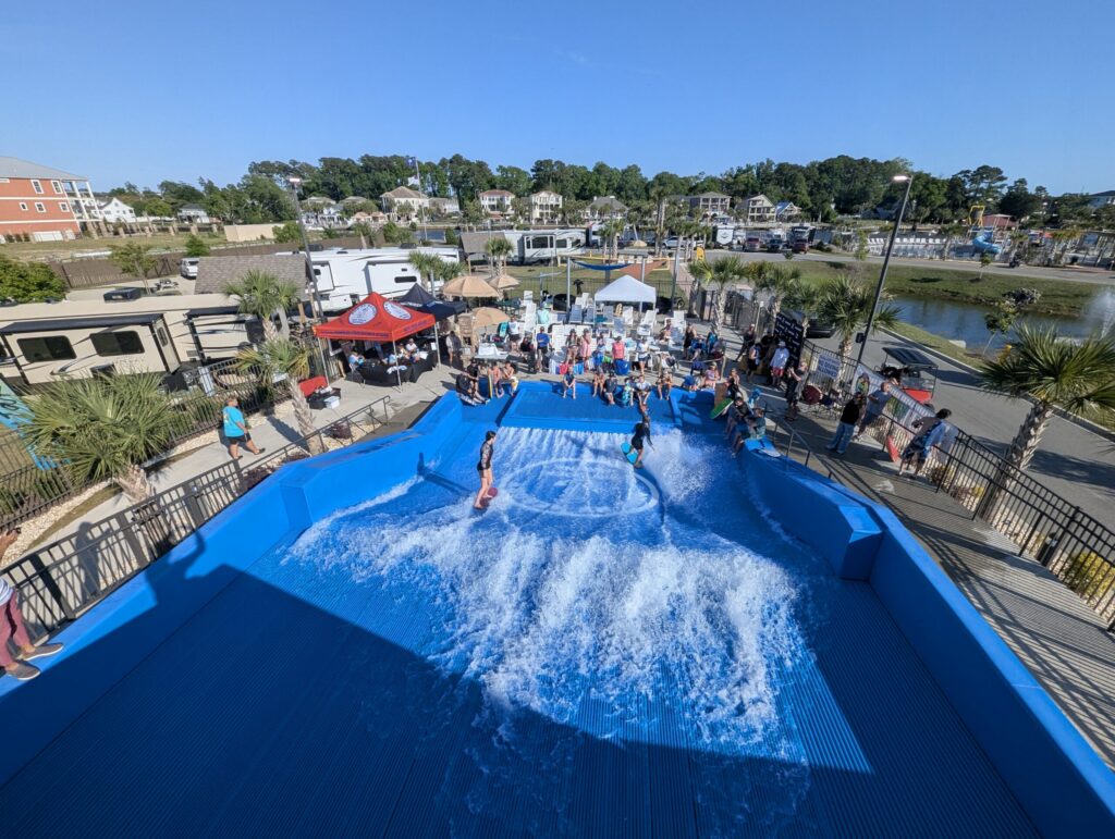 People watch as several children and teens ride artificial waves on a blue Flow Tour surfing simulator at an outdoor recreational area, with palm trees, RVs, and buildings in the background under a clear sky.