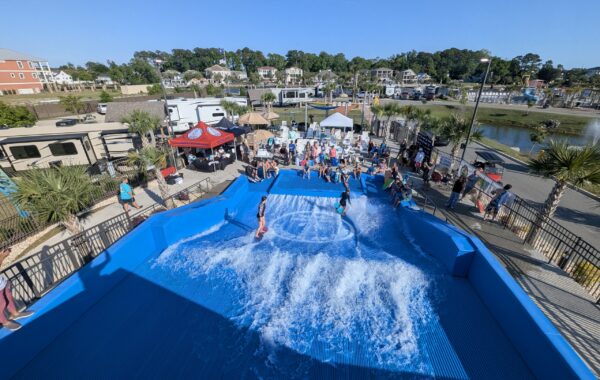 People watch as several children and teens ride artificial waves on a blue Flow Tour surfing simulator at an outdoor recreational area, with palm trees, RVs, and buildings in the background under a clear sky.