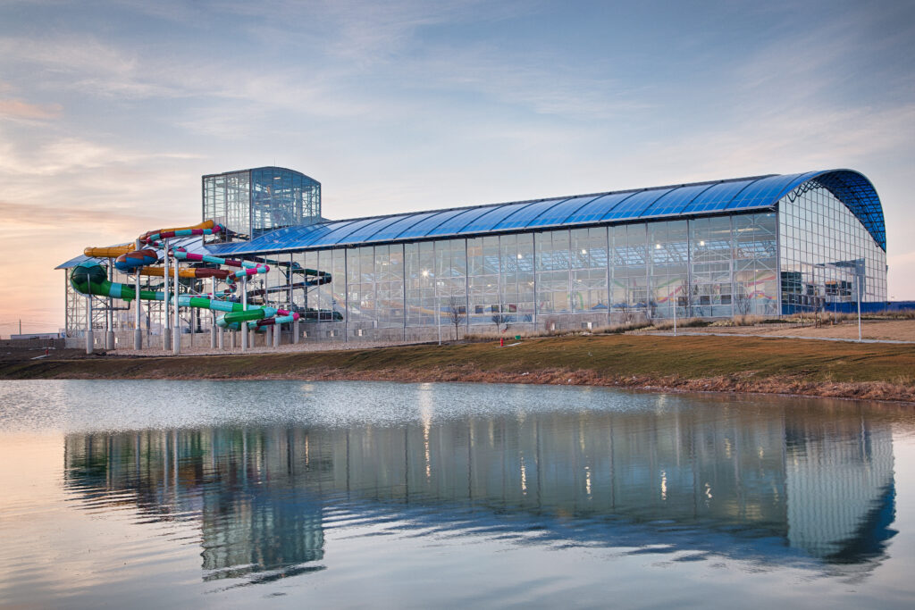 A large glass-covered indoor water park featuring colorful FlowSurf water slides is situated beside a calm pond, reflecting the building and slides at sunset.