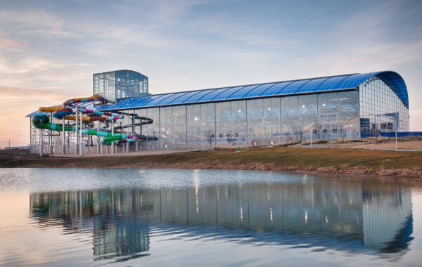 A large glass-covered indoor water park featuring colorful FlowSurf water slides is situated beside a calm pond, reflecting the building and slides at sunset.
