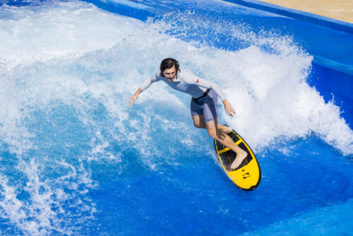 A man in a light gray rash guard and blue shorts rides a yellow surfboard on an artificial wave during the Flow Tour at a water park, surrounded by splashing blue water.