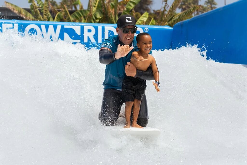 An adult and a young child surf together on an artificial wave, both smiling. The adult supports the child, who stands confidently at the front of the surfboard. Splashes of water surround them.