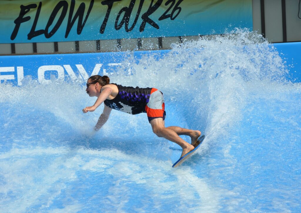 A person in a black and purple tank top and red shorts rides a flowboard on an artificial wave during the Flow Tour, kicking up water as they balance with knees bent and one hand extended. A “FLOW TOUR” banner is visible in the background.