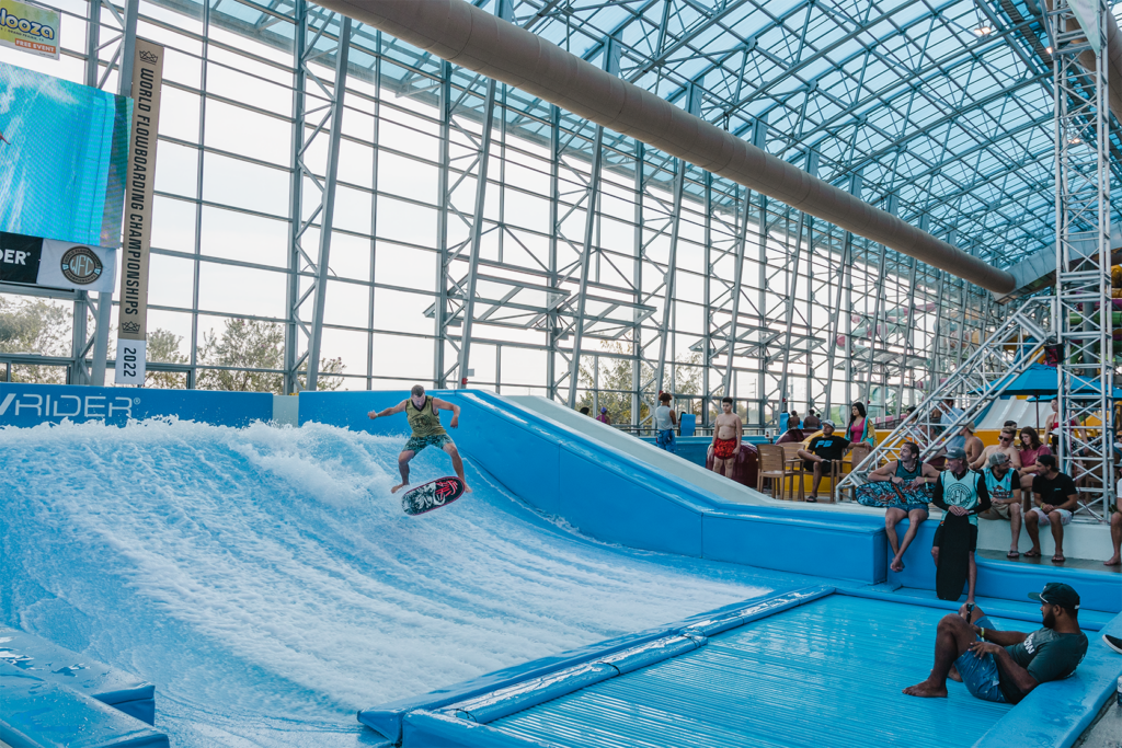 A person rides a wave on the FlowSurf indoor surfing simulator while several people watch from seats nearby. The setting is bright, featuring a large glass ceiling and sleek metal structures.