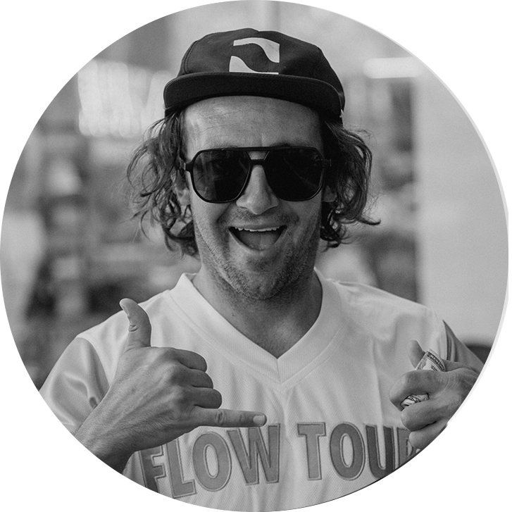A man wearing sunglasses, a cap, and a FLOW TOUR shirt smiles and makes a shaka hand gesture with both hands. The image is black and white and taken in a circular frame.