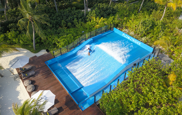 A person surfs on a blue FlowSurf wave simulator surrounded by lush green vegetation, palm trees, and white sand, with lounge chairs and umbrellas nearby.