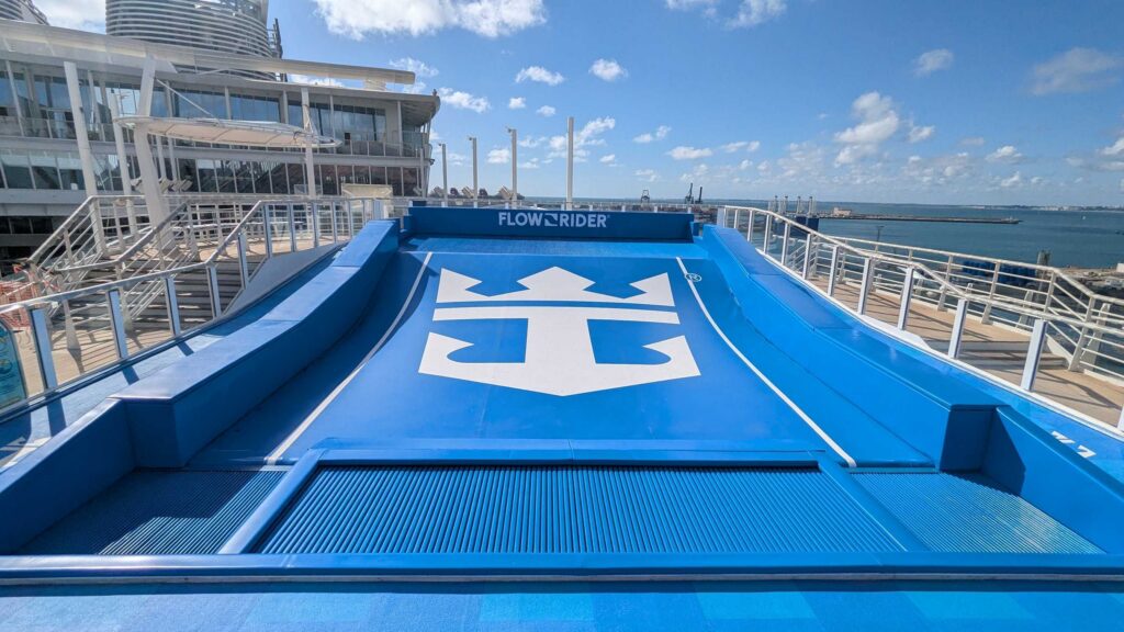 A blue FlowRider and FlowSurf surf simulator with the Royal Caribbean logo sits on a cruise ship deck, surrounded by railings and overlooking the ocean under a partly cloudy sky.